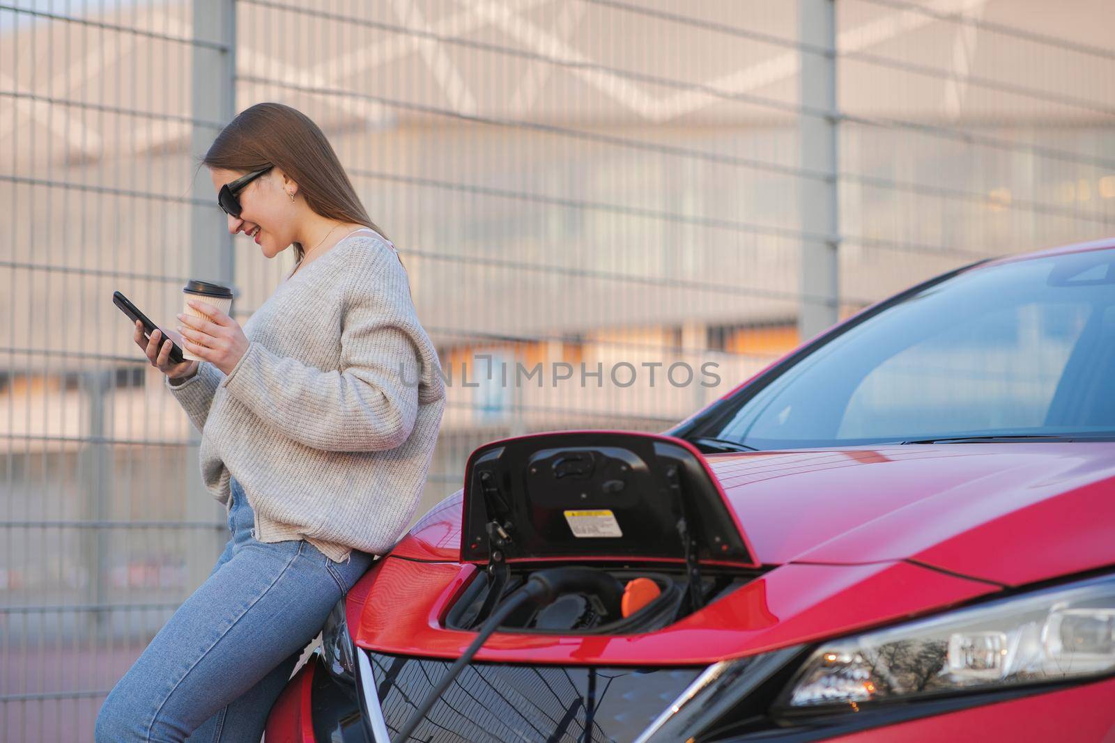 Electric car charging in street. Ecological Car Connected and Charging Batteries. Girl Use Coffee Drink While Using SmartPhone and Waiting Power Supply Connect to Electric Vehicles for Charging.