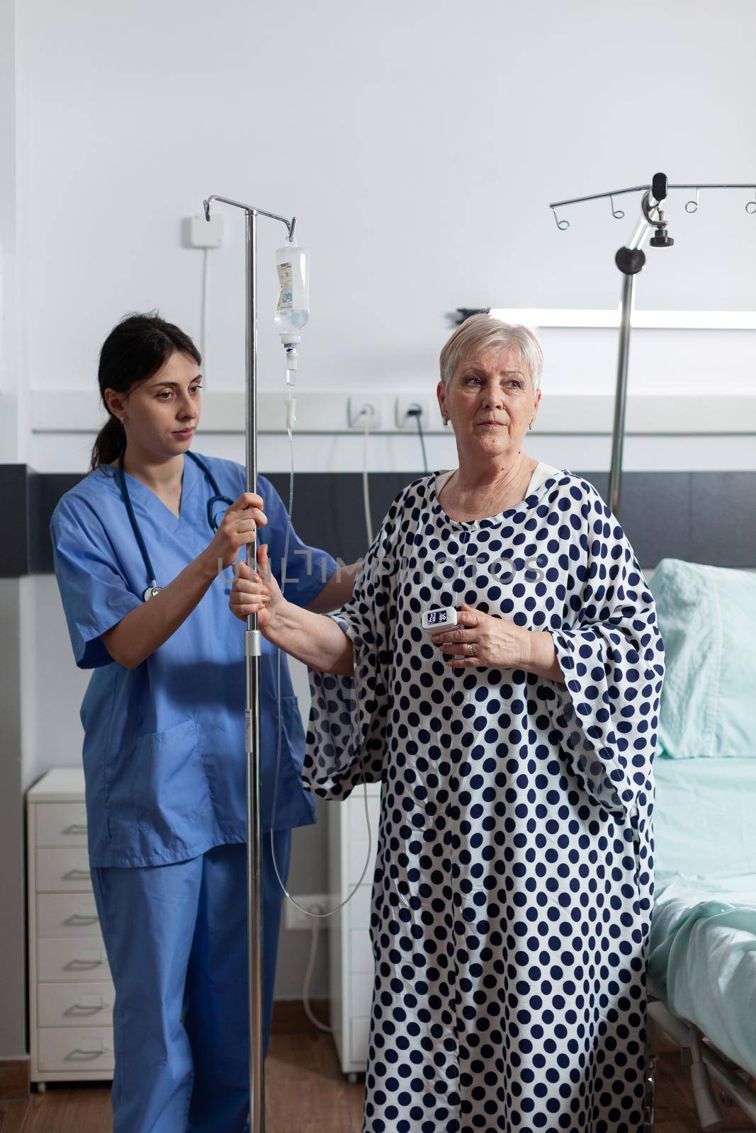 Nurse dressed with scrubs helping senior woman patient with iv drip bag attached while getting intravenous medicine. Pensioner with oxymeter on finger.