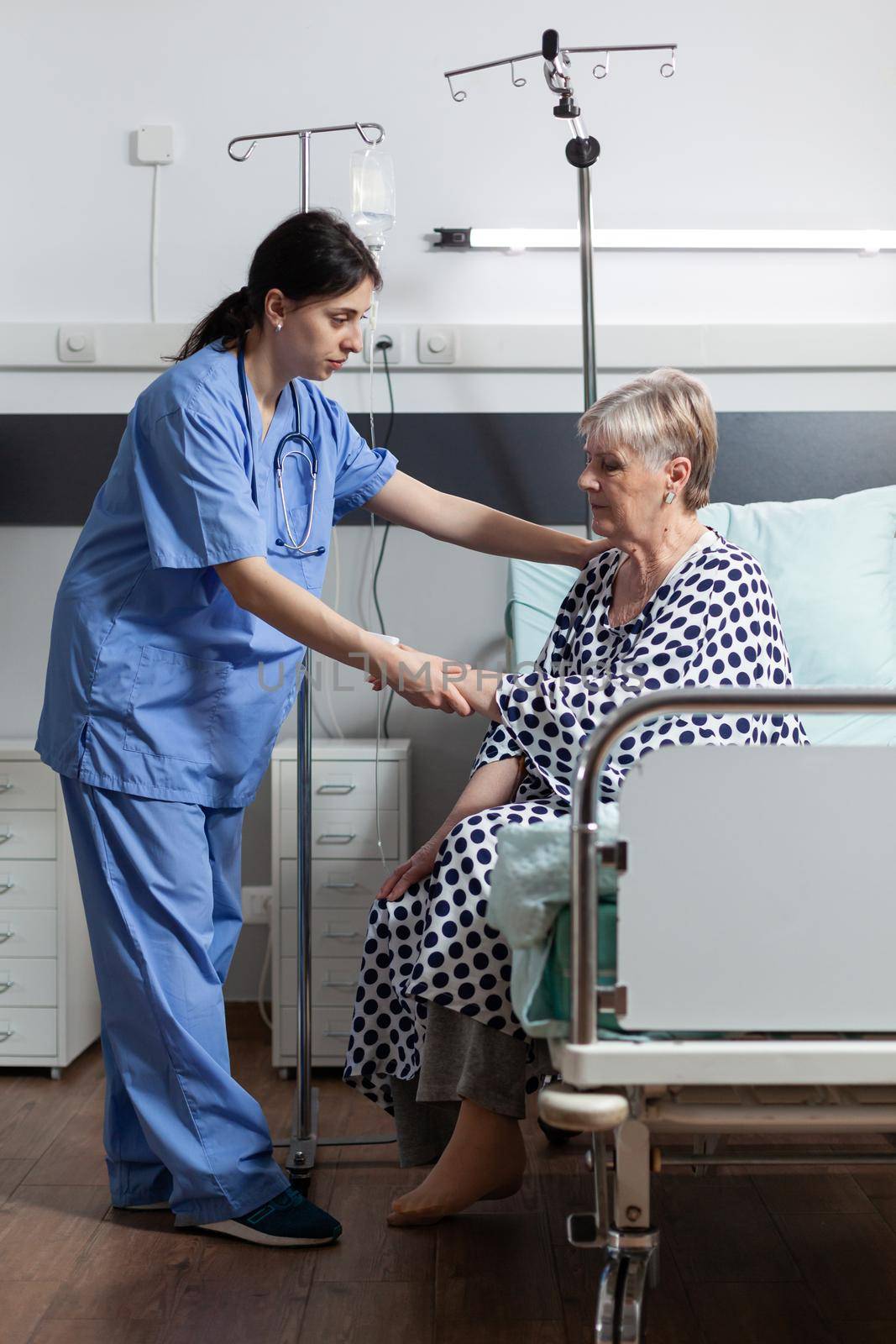 Medical nurse helping senior woman patient getting up from bed in hospital room, with iv drip bag attached while she gets intravenous treatment and oxymeter measuring blood oxygen saturation.