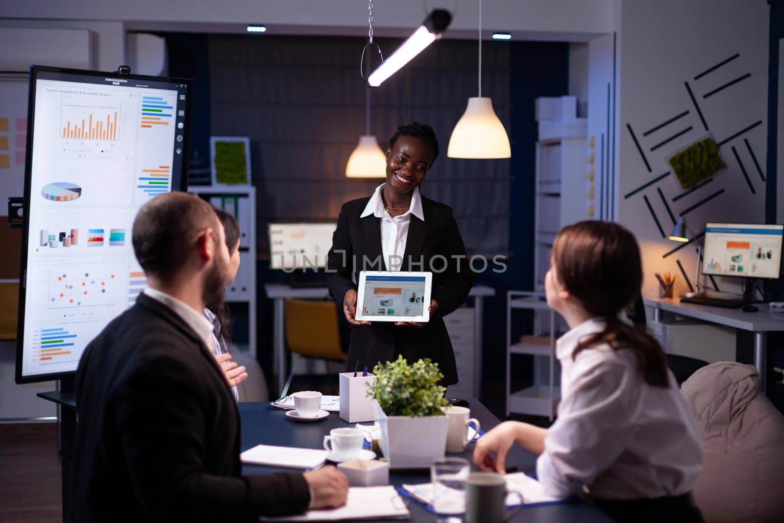 Focused workaholic entrepreneur woman with dark skin explaining management strategy using tablet. Business diverse multi-ethnic teamwork overworking in company office meeting room late at night