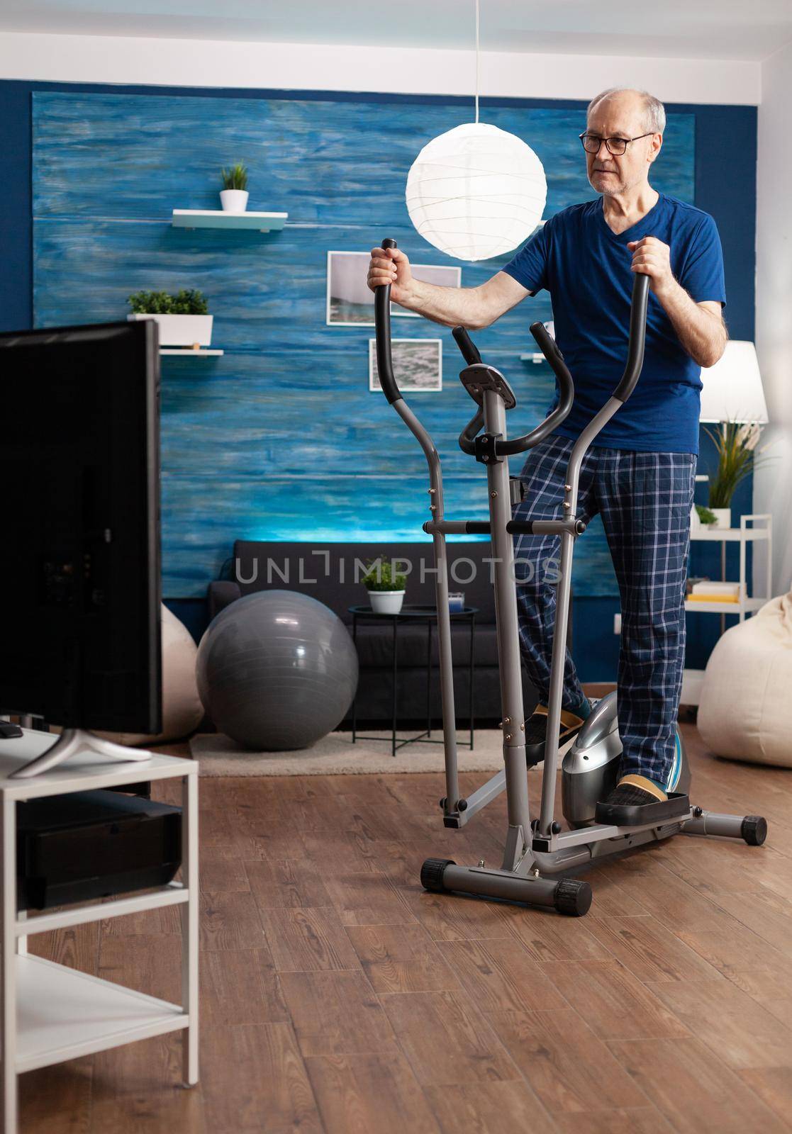Focused retirement senior man working at legs resistance using cycling bicycle machine watching aerobics video on television for well being. Pensioner training body muscle during health cardio workout