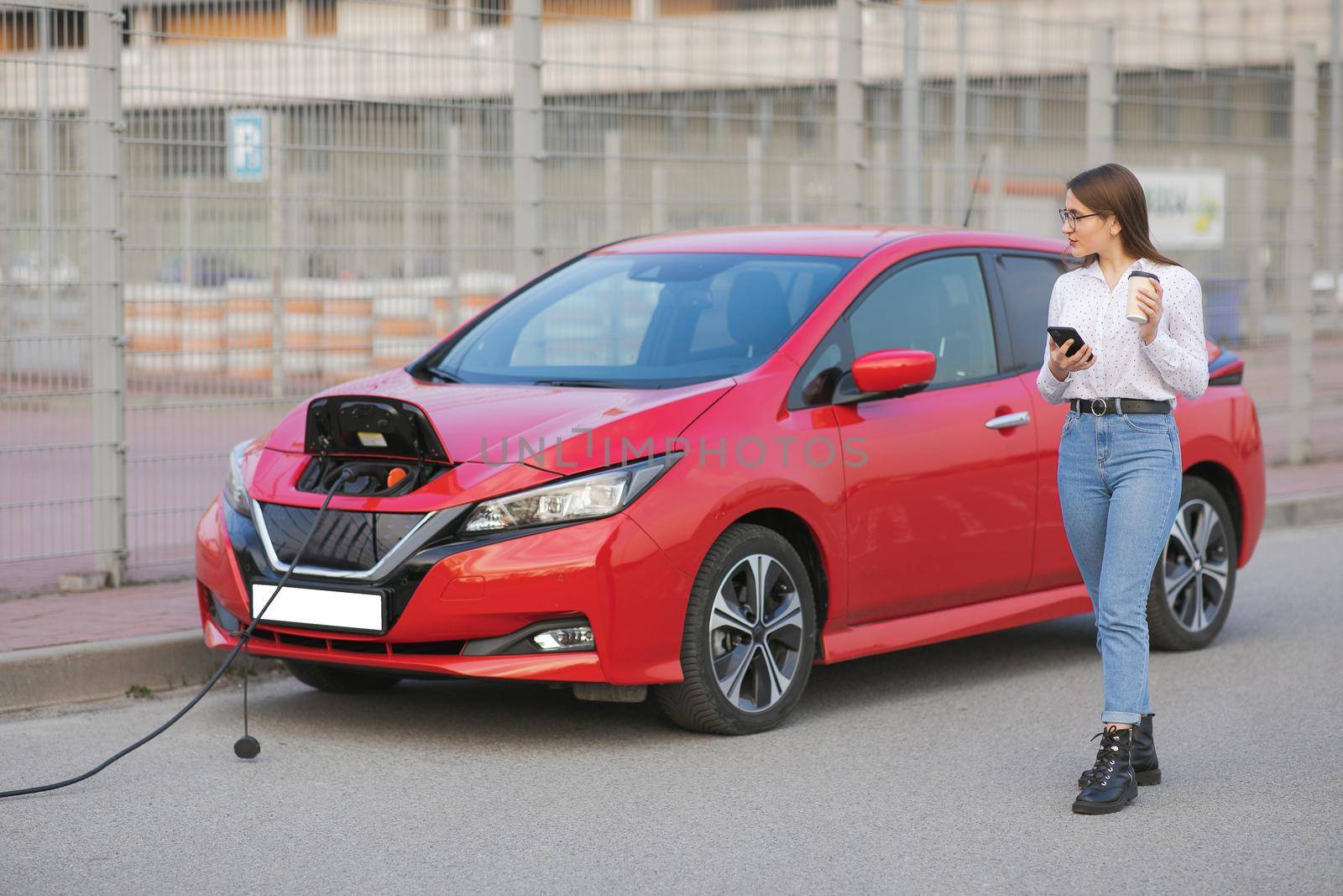 Girl Use Coffee Drink While Using Smart Phone and Waiting Power Supply Connect to Electric Vehicles for Charging the Battery in Car. by uflypro