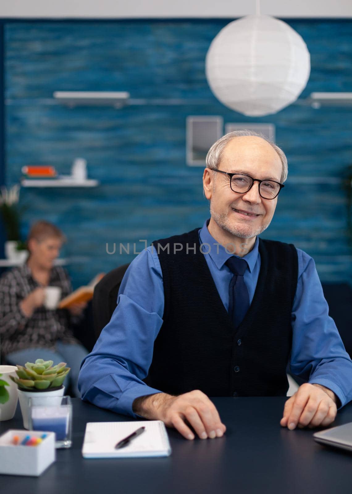 Cheerful senior businessman with gray hair. smiling at camera Elderly man entrepreneur in home workplace using portable computer sitting at desk while wife is reading a book sitting on sofa.