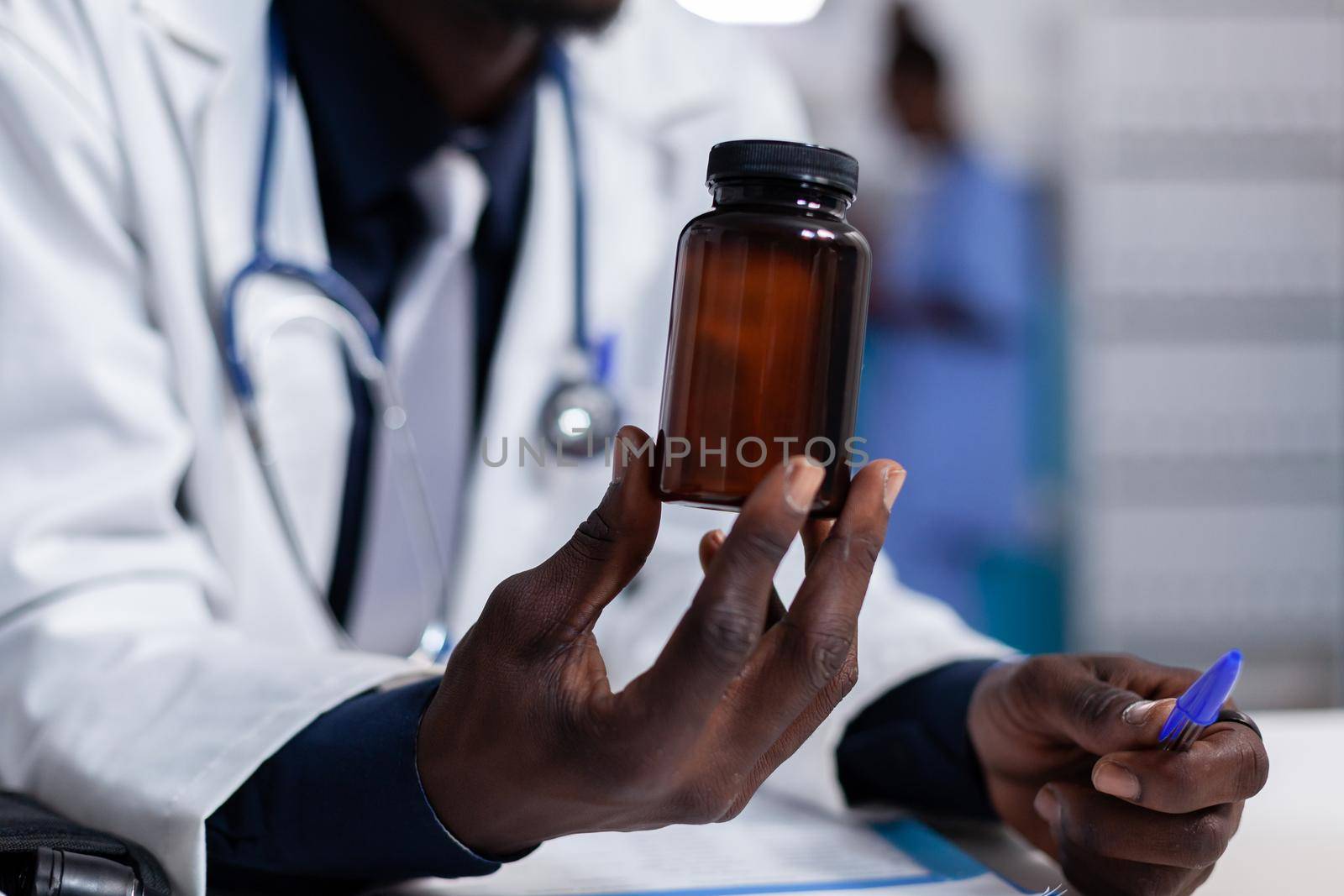 Close up of african american hand with bottle of pills for treatment recommendation. Black doctor holding flask with medicine, medicament and drugs. Medic with vial in medical cabinet