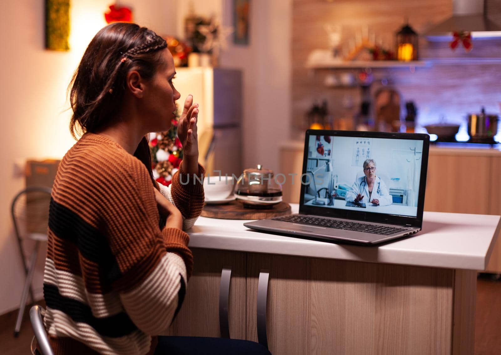 Woman with sickness calling doctor on online video conference while sitting in festive kitchen at home. Ill young patient using telemedicine for healing treatment and healthcare advice