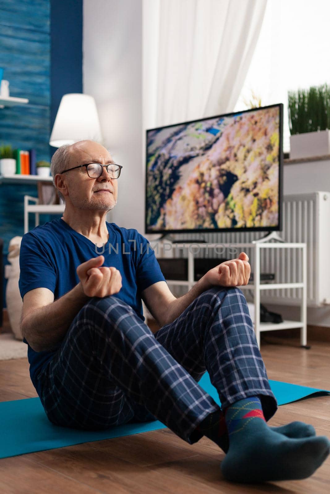 Peaceful senior man practicing meditation workout sitting comfortable in lotus position with closed eyes on yoga mat. Caucasian male working at body exercise posture in living room