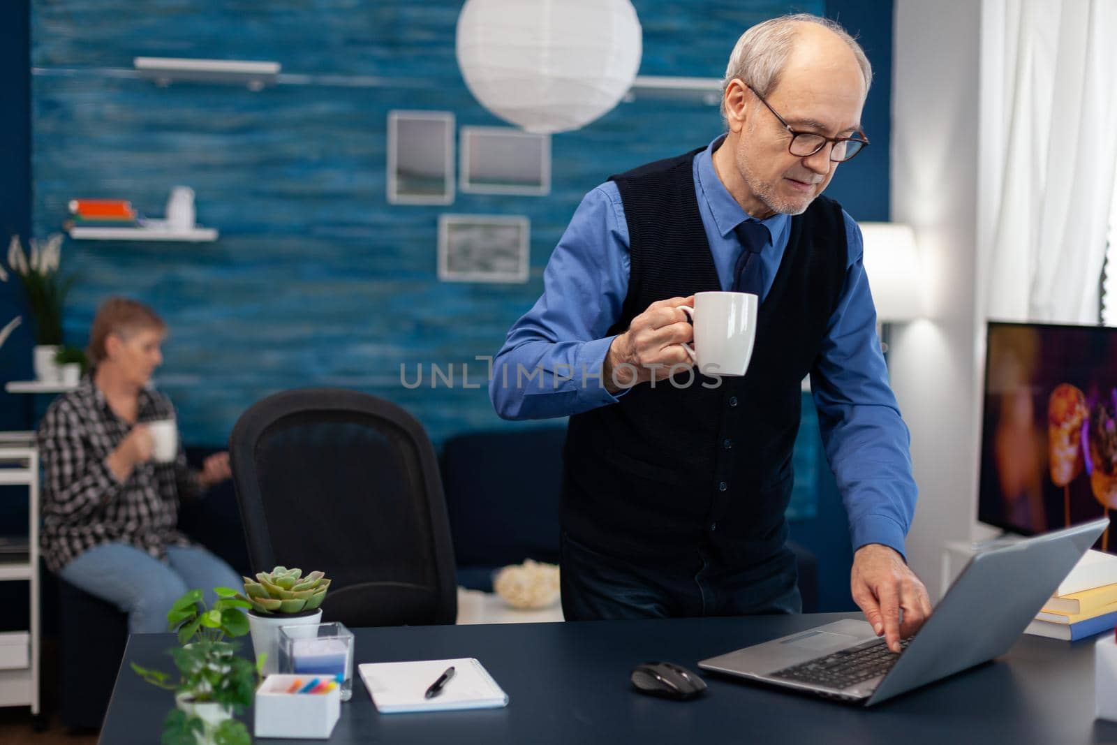 Retired businessman turning on laptop enjoying a cup of coffee. Elderly man entrepreneur in home workplace using portable computer sitting at desk while wife is holding tv remote.