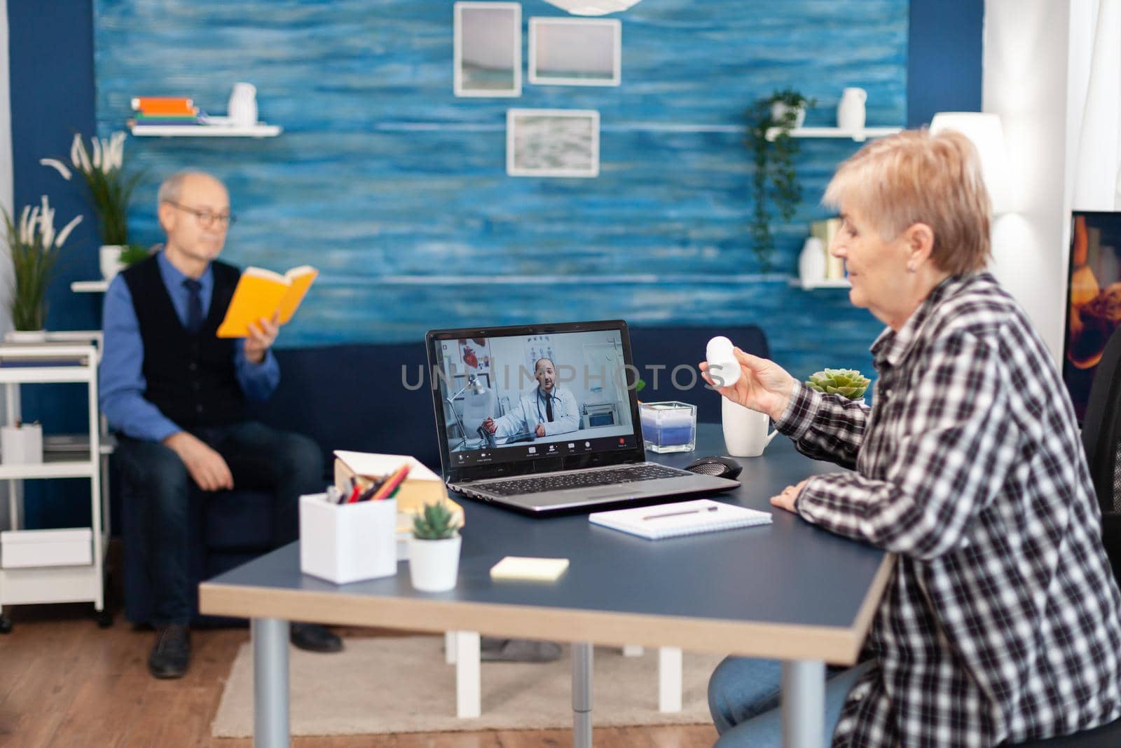 Older woman listening doctor indication about health treatment during call. Elderly woman discussing with healthcare practitioner in the course of remote call and husband is reading a book on sofa.