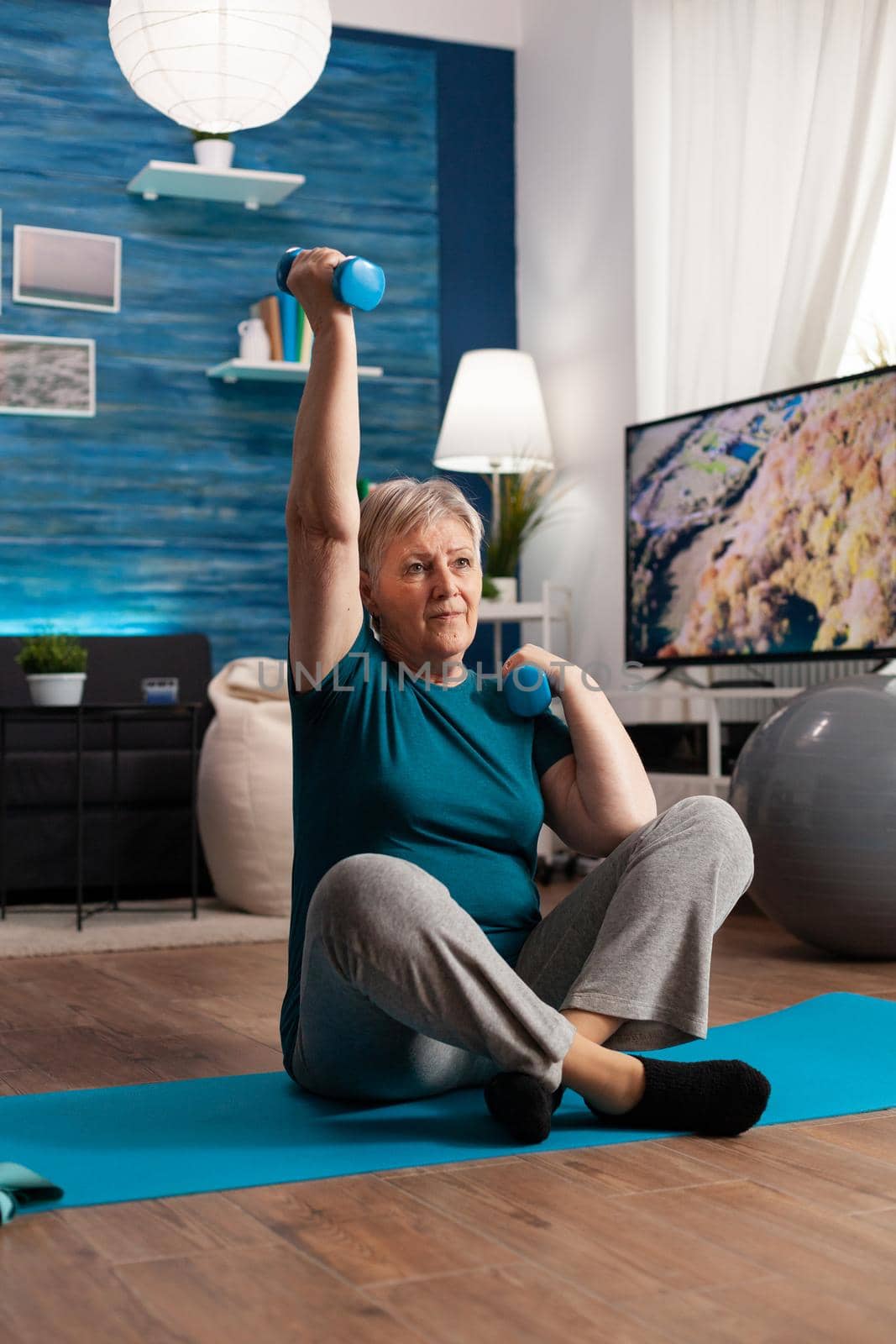 Retired senior woman sitting on yoga mat in lotus position raising hand during wellness routine warming up training body muscles using dumbbells. Pensioner in sportswear practicing abs exercise