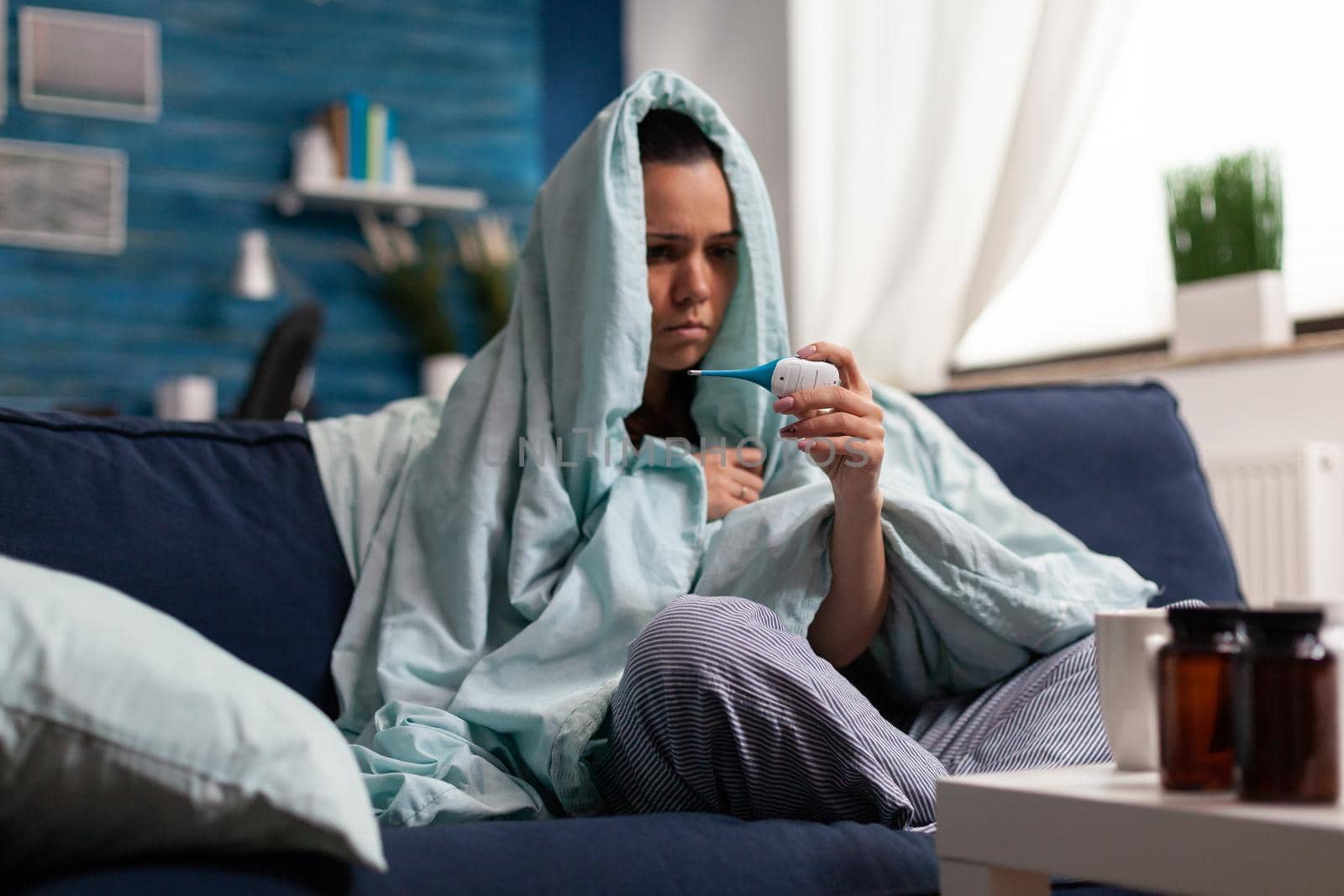 Young woman with flu symptoms checking temperature at home using thermometer. Adult measuring fever sitting on sofa in blanket. Tired unhealthy human in pain taking medical treatment