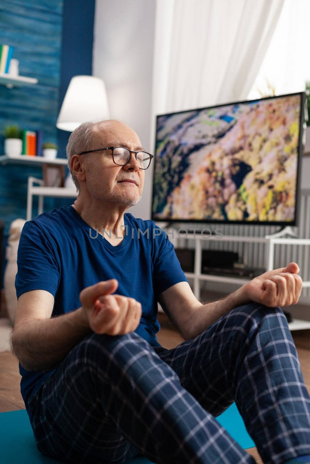 Healthy senior man sitting comfortable in lotus position on yoga mat with closed eyes by DCStudio