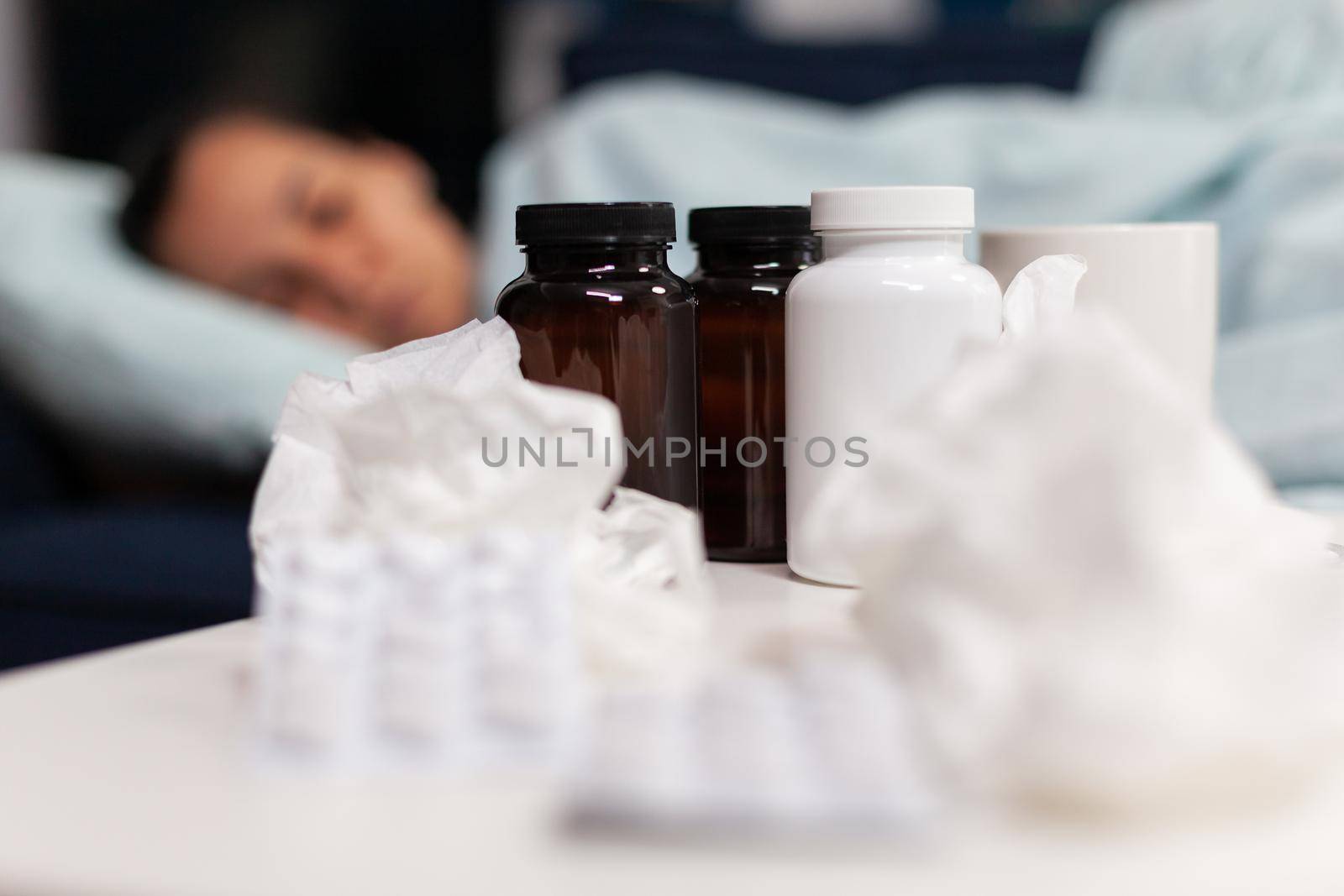 Close up of medication, pills and napkins for sick woman sleeping on sofa. Taking drugs and medical treatment against cold flu virus symptoms. Young adult with infection and temperature.
