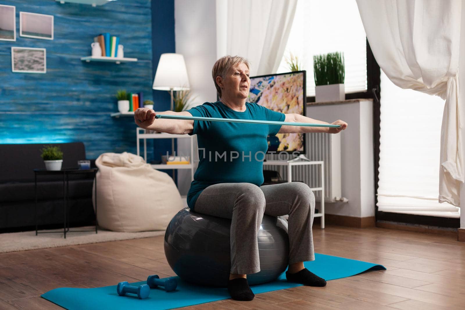 Retired pensioner sitting on swiss ball working at body weight stretching muscle arm using fitness elastic band. Active senior woman watching online sport video on laptop practicing strech exercise