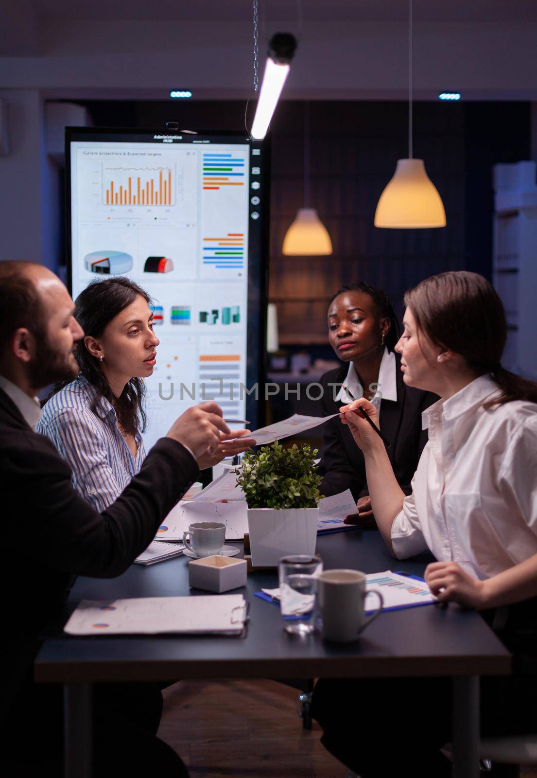 Businesswoman leder sitting at desk analyzing marketing presentation discussing financial graps paperwork. Diverse multi-ethnic teamwork brainstorming project ideas working in meeting room late night
