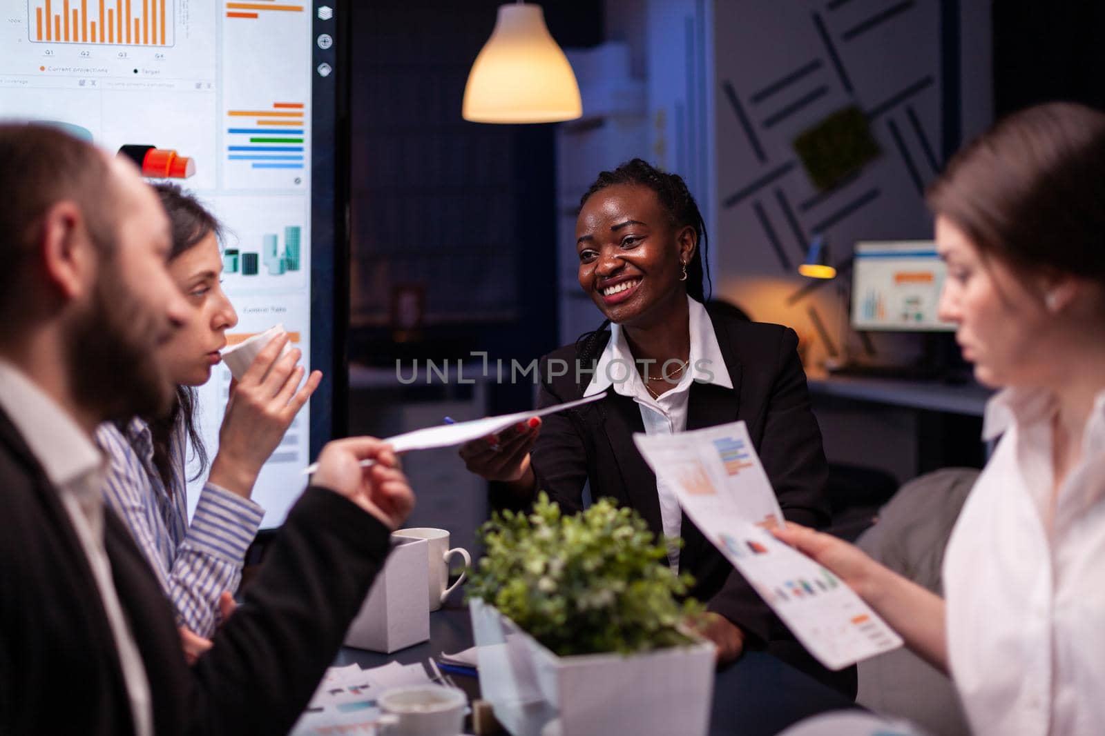 Workaholics focused multi-ethnic businesspeople overworking in company office meeting room brainstorming ideas late at night. Coworkers sitting at desk analyzing statistics paperwork