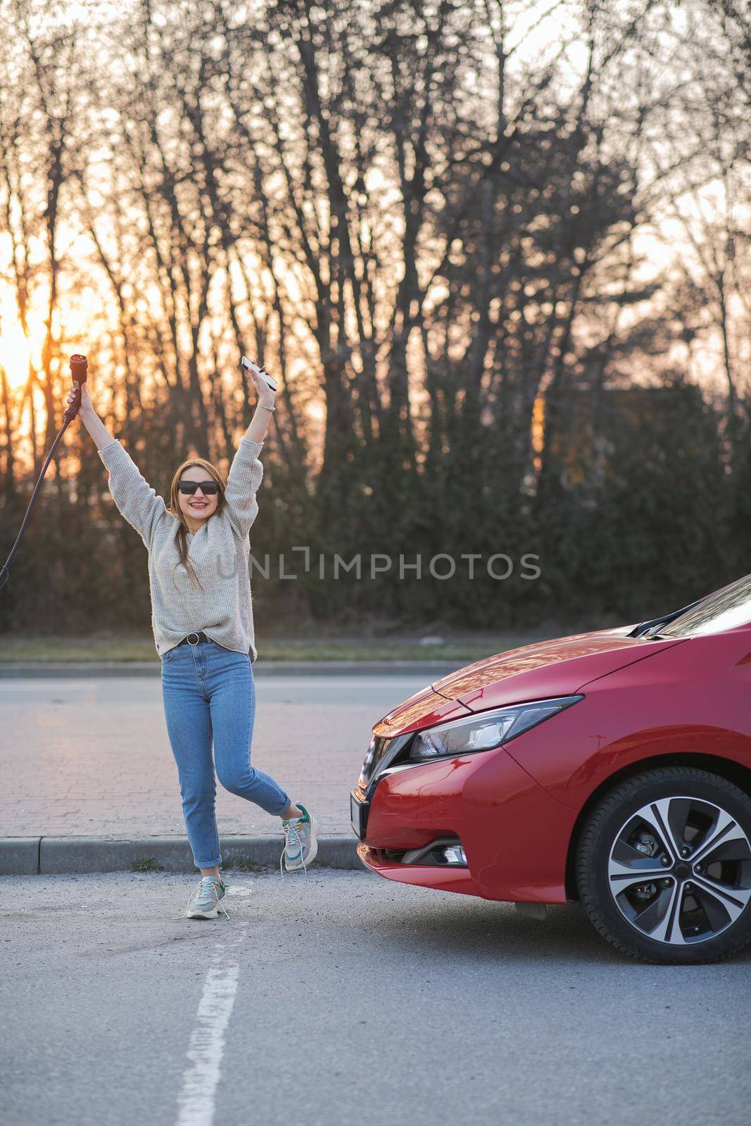 Happy woman charging electric car on charging station, focus on charger. Beautiful woman with electric plug in her hand goes to the car for charging