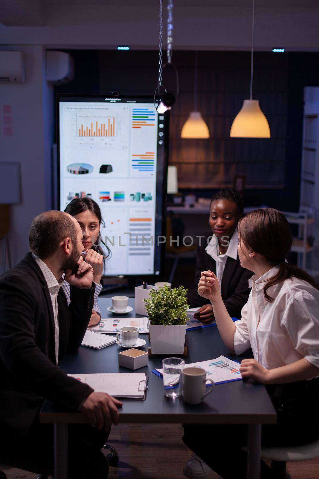 Workaholics businesspeople brainstorming financial company ideas analyzing strategy paperwork late at night in business office meeting room. Multi-ethnic coworkers checking solution
