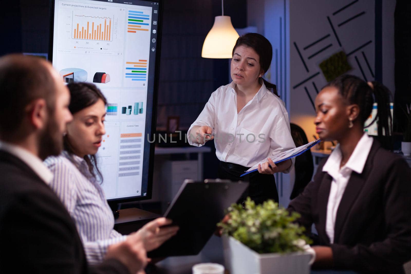 Diverse multi ethnic teamwork brainstorming company ideas analyzing financial graphs presentation. Businesspeople overworking in meeting room late at night discussing solution