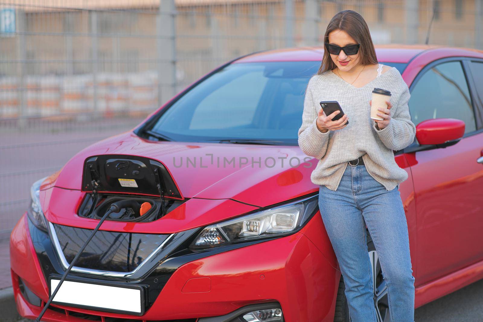 Woman stands with phone near her electric car and waits when vehicle will charged. Charging of electric car.
