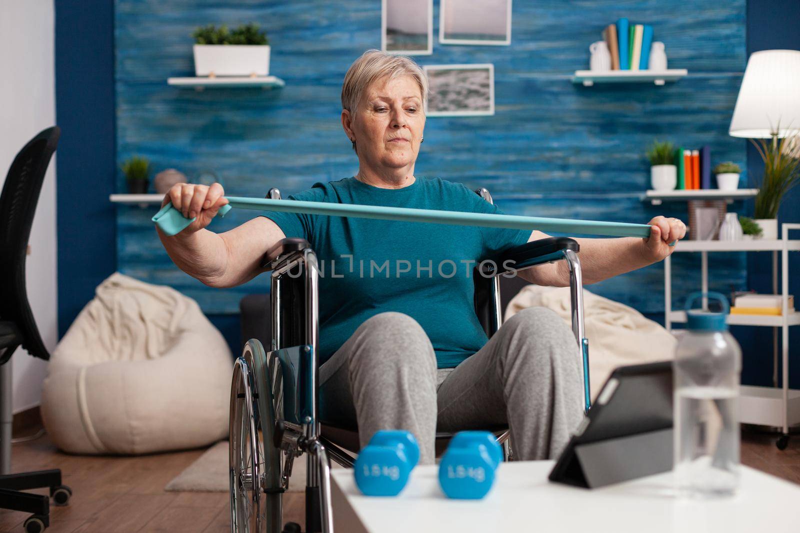 Invalid senior woman exercising arm muscle resistance using elastic band watching fitness video on tablet doing recovery aerobics exercise. Pensioner working at body healthcare therapy in living room