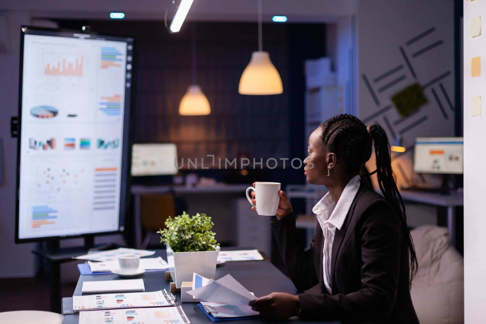Workaholic african american businesswoman analysing marketing statistics on paperwork drinking coffee sitting at table late at night. Business manager with dark skin working in office meeting room
