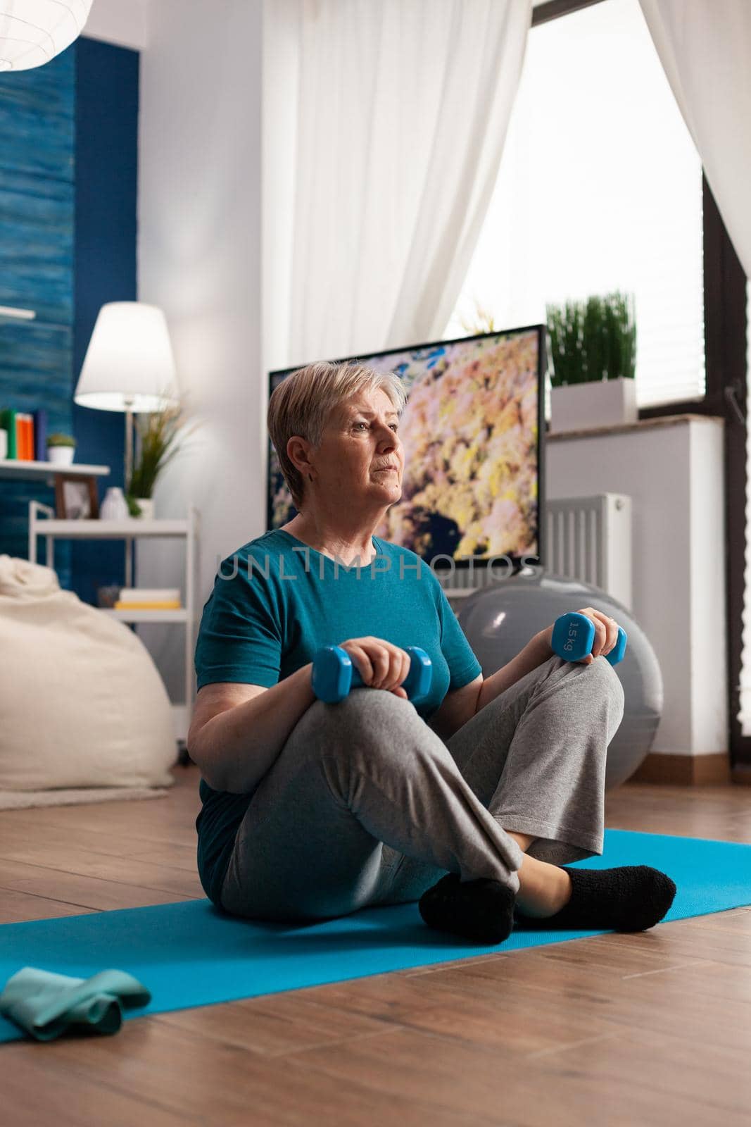 Retired senior woman sitting on yoga mat holding fitness dumbbells in lotus position during pilates wellness routine. Healthy pensioner training abdominals muscles practicing pilates in living room
