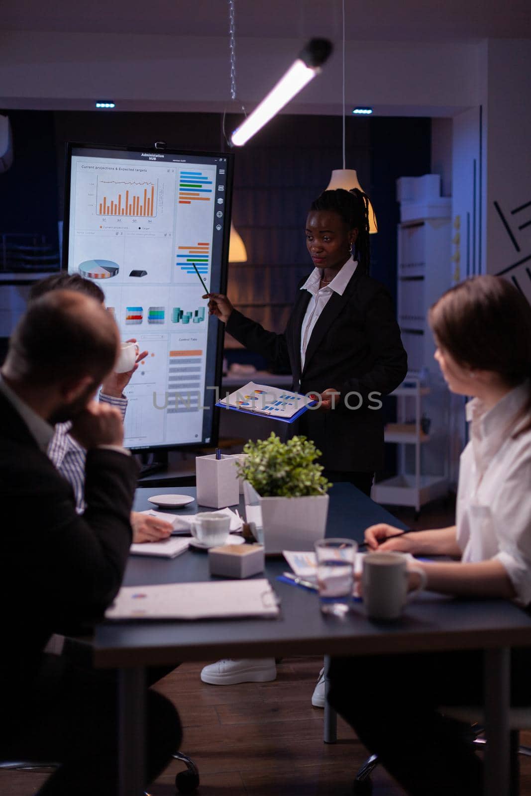 Businesswoman with dark skin explaining financial graphs using presentation monitor overworking in company meeting room in evening. Workaholics multi-ethnic teamwork solving statistics at night