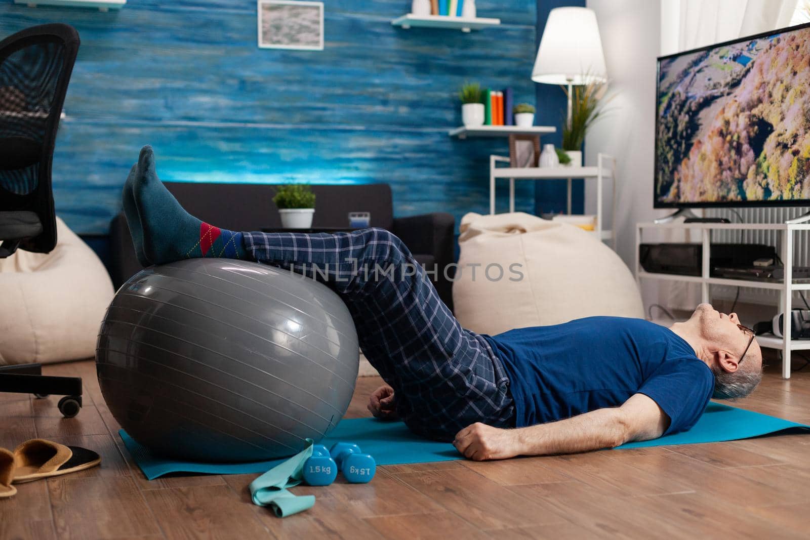 Retirement senior man doing wellness warming legs up using swiss ball sitting on yoga mat in living room. Retired pensioner practicing abdominals exercises working at body muscular healthcare