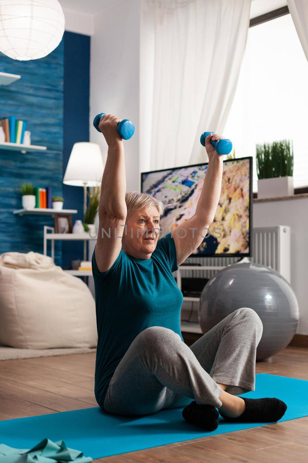 Retired senior woman sitting on yoga mat in lotus position raising hands streching arm muscle doing fitness exercise using dumbbells during wellness workout. Athlete pensioner slimming weight