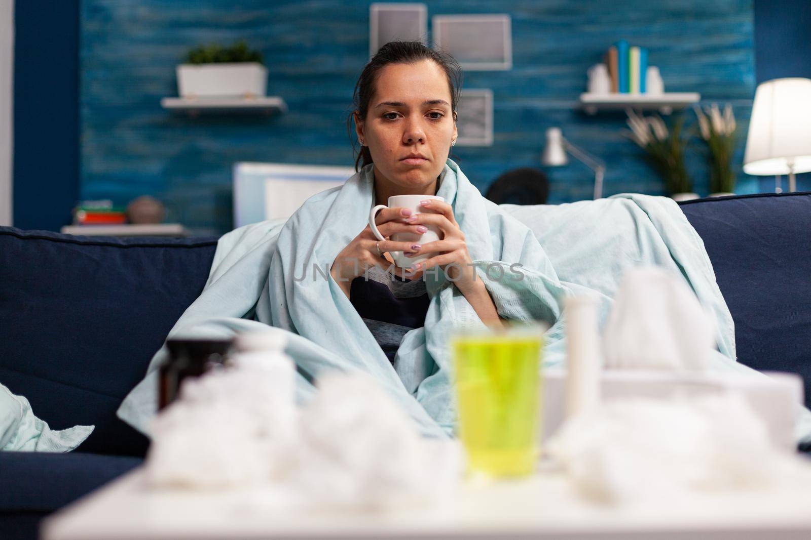 Sick young adult with seasonal infection sitting at home wrapped in blanket, drinking hot tea against flu fever disease. Mug of warm drink as medicine to treat virus symptoms