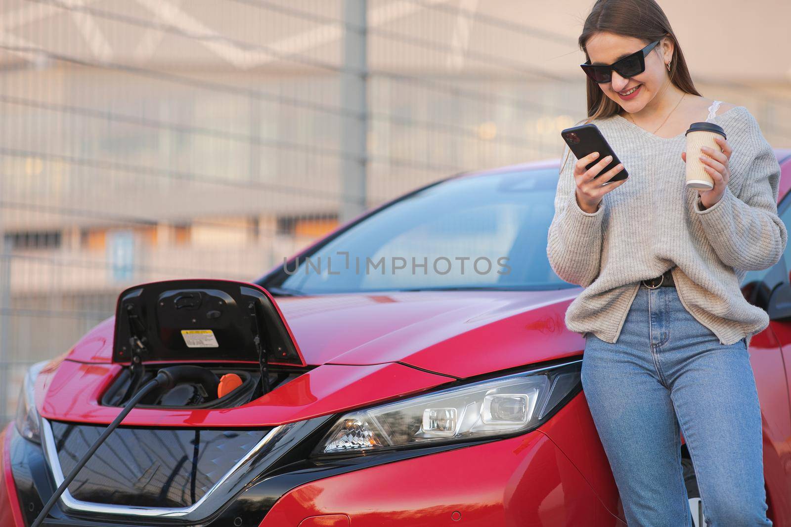 Girl Use Coffee Drink While Using Smart Phone and Waiting Power Supply Connect to Electric Vehicles for Charging the Battery in Car