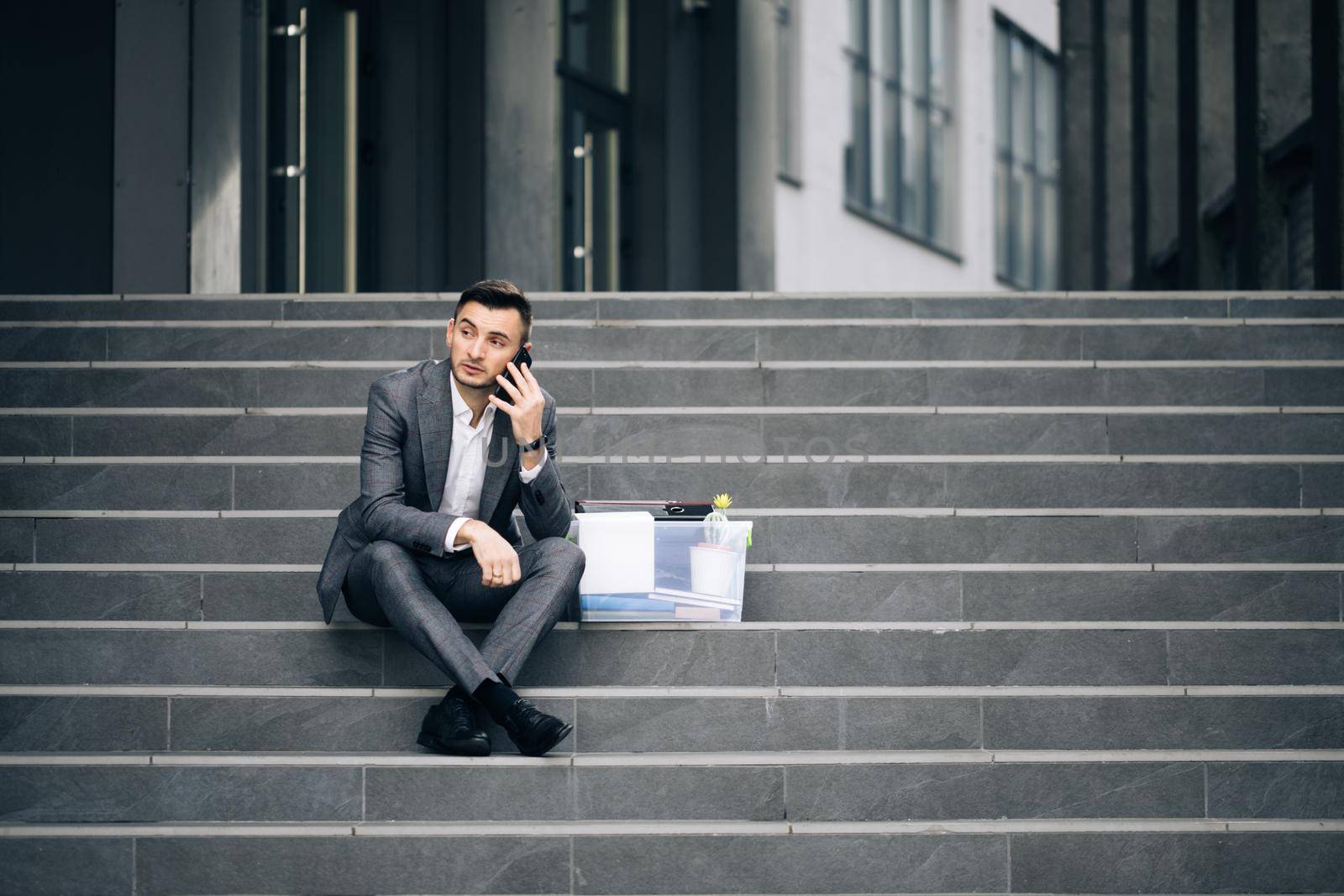 Young male jobless office worker in suit sitting on stairs outdoors. Fired man talking on phone with stuff in box. Cellphone conversation. Speaking on telephone by uflypro
