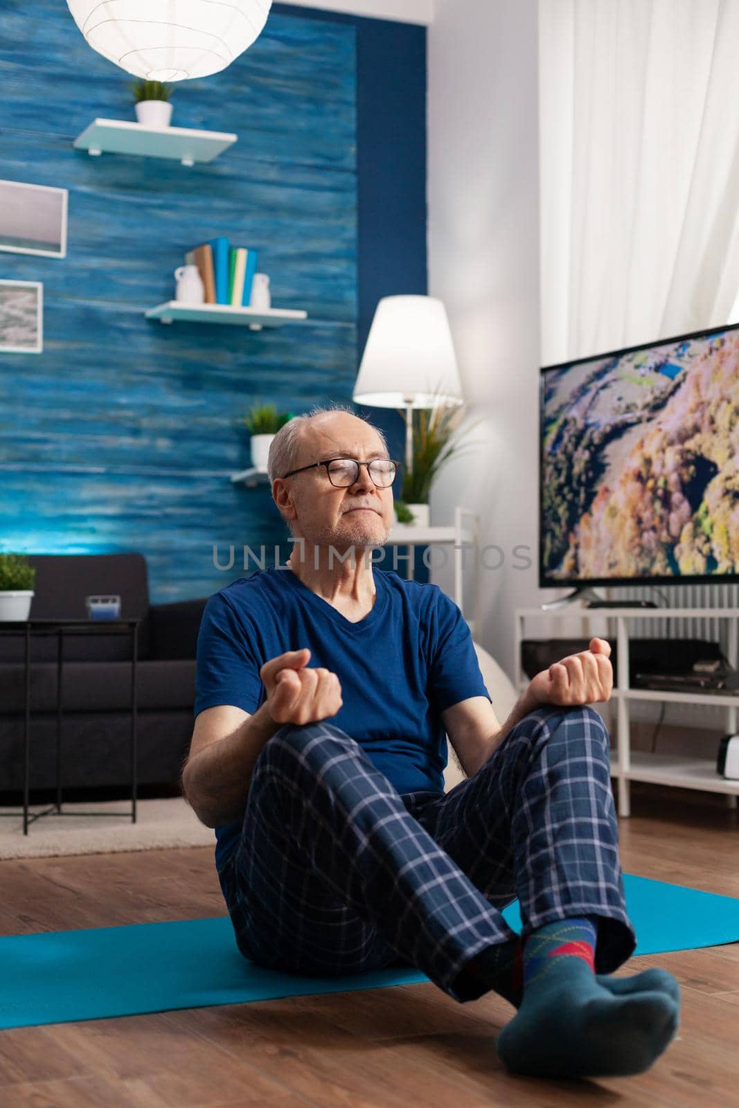 Senior man sitting comfortable in lotus position on yoga mat with closed eyes training body posture. Peaceful pensioner practicing fitness exercise during meditation workout in living room