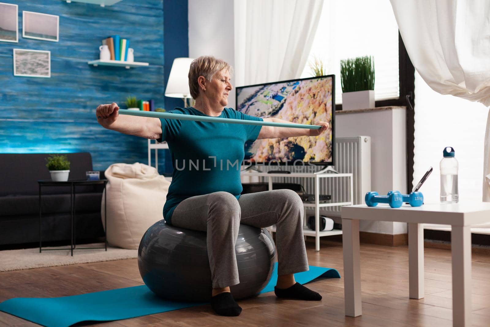 Cheerful retired pensioner working arm muscles using elastic band practicing aerobic exercise. Pensioner sitting on swiss ball in living room working at body healthcare resistance watching gym video