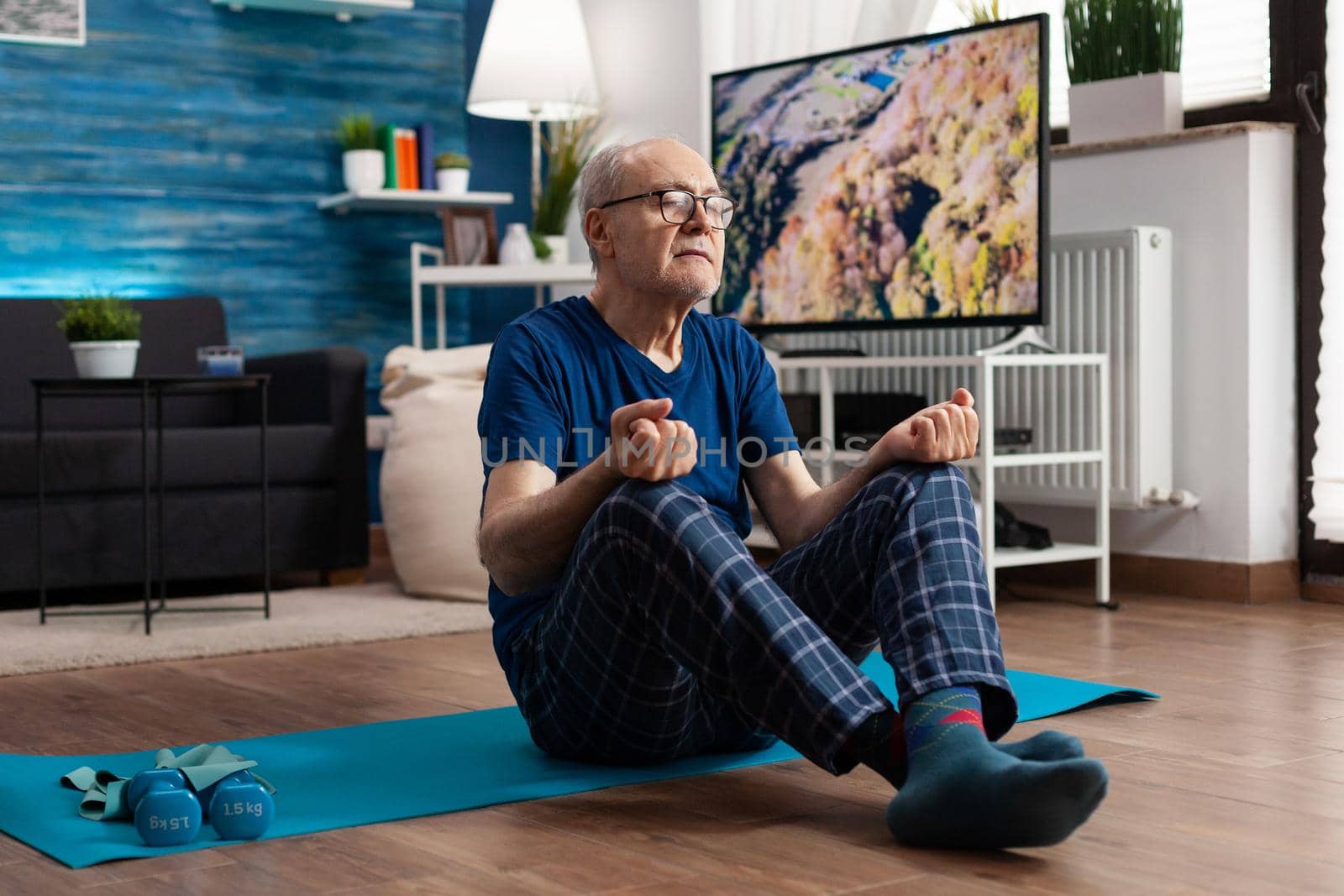 Retreat senior man sitting comfortable in lotus position on yoga mat with closed eyes doing pilates. Peaceful pensioner practicing fitness body exercise during meditation workout in living room