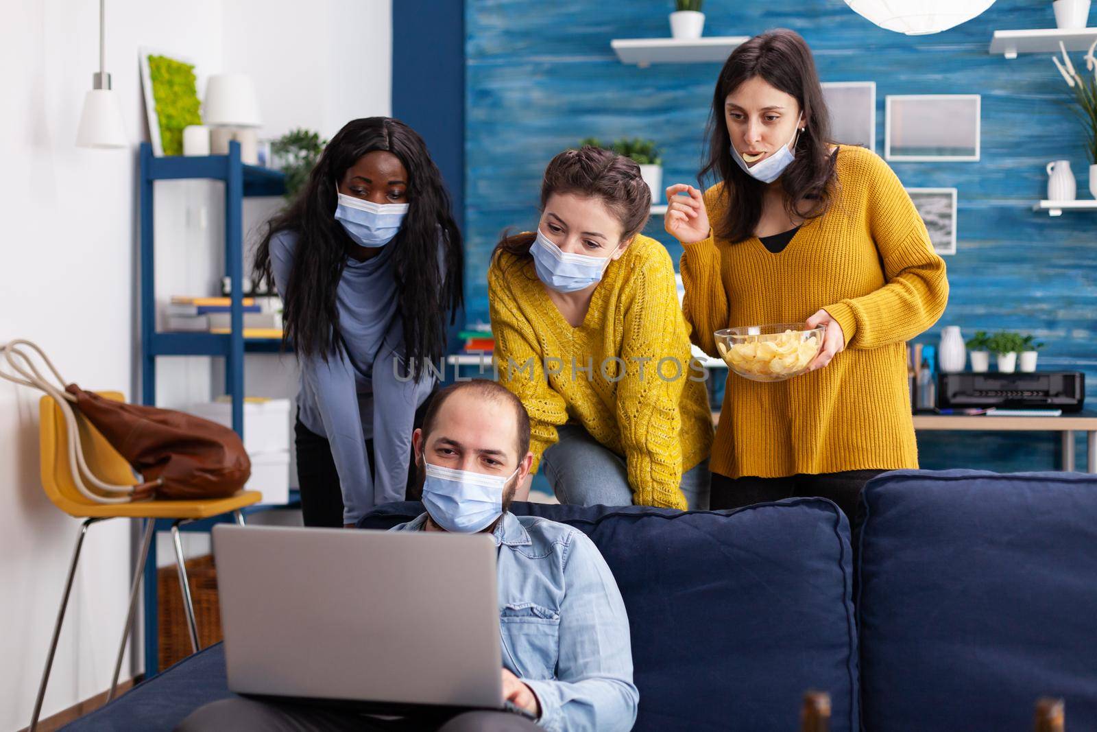 Group of multi ethnic friends keeping social distancing wearing face mask to prevent spread of coronavirus looking at laptop in home living room spending time together drinking beer eating chips. Conceptual image.