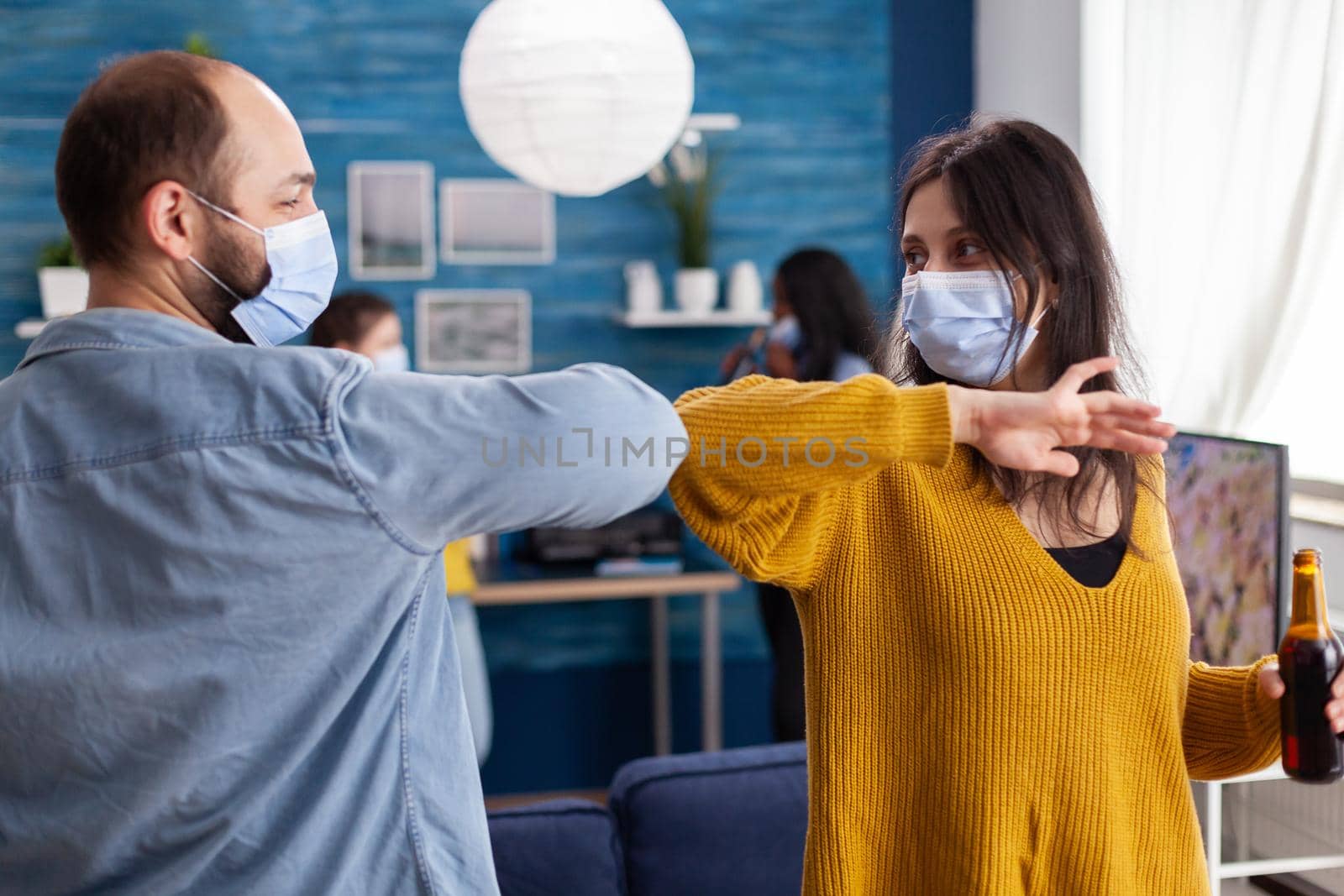 Happy cheerful friends touching elbow keeping social distancing greeting eachother wearing face mask preventing spread of coronavirus holding beer bottles in apartment living room. Conceptual image.