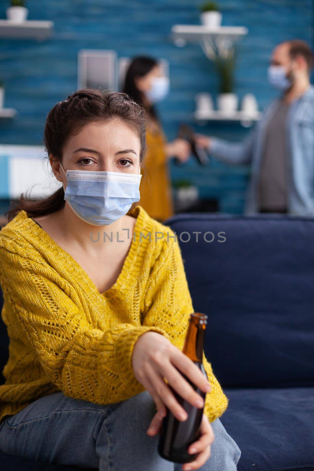 Happy woman and friends spending time together in apartment living room talking keeping social distancing wearing face mask as prevention for coronavirus spreading in the course of global pandemic.