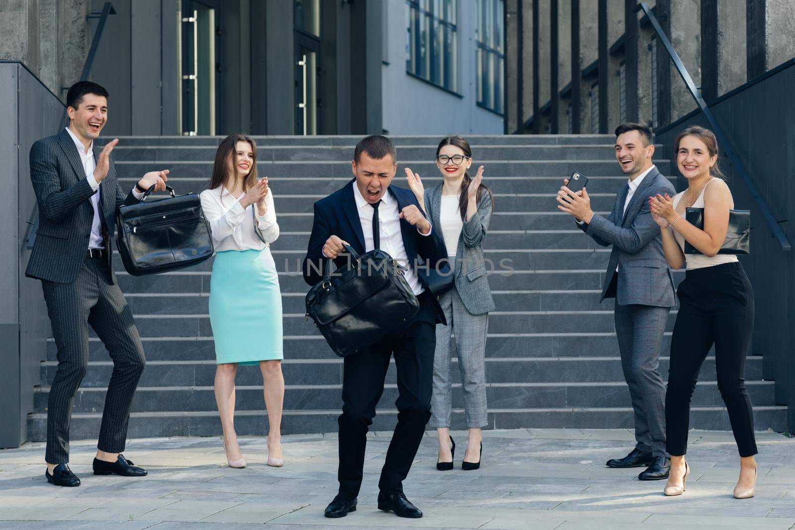 Young Happy Business Manager Wearing a Suit and Tie Dancing from Office Building. Colleagues are Cheering. Diverse and Motivated Business People from Modern Office by uflypro