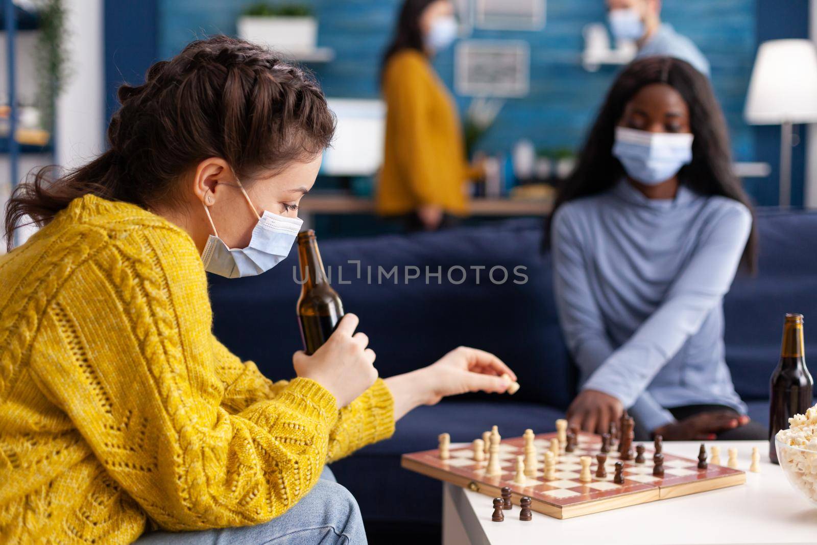 Multiethnic friends playing chess in home living room chatting talking keeping social distancing to prevent illness with covid19 during global pandemic wearing face mask. Conceptual image. Board games, competing, tactics, asctivity.