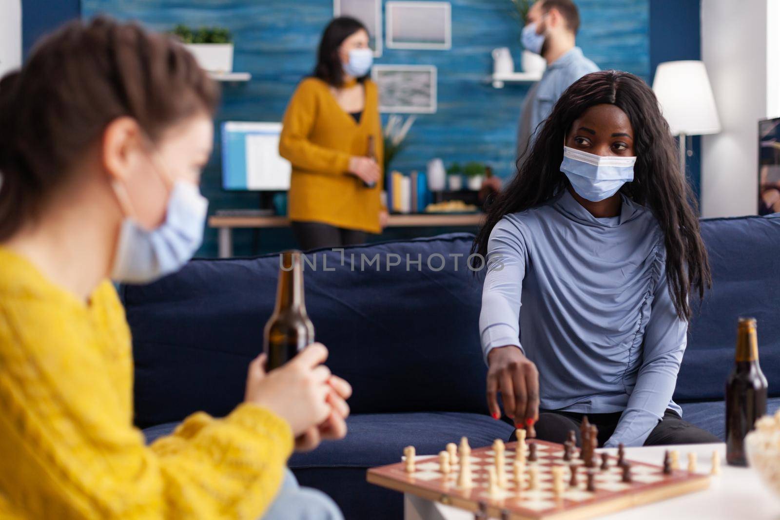 African woman wearing face mask playing chess with friends keeping social distancing chatting aving fun, drinking beer preventing spread of covid virus. Board games, competing, tactics, asctivity.