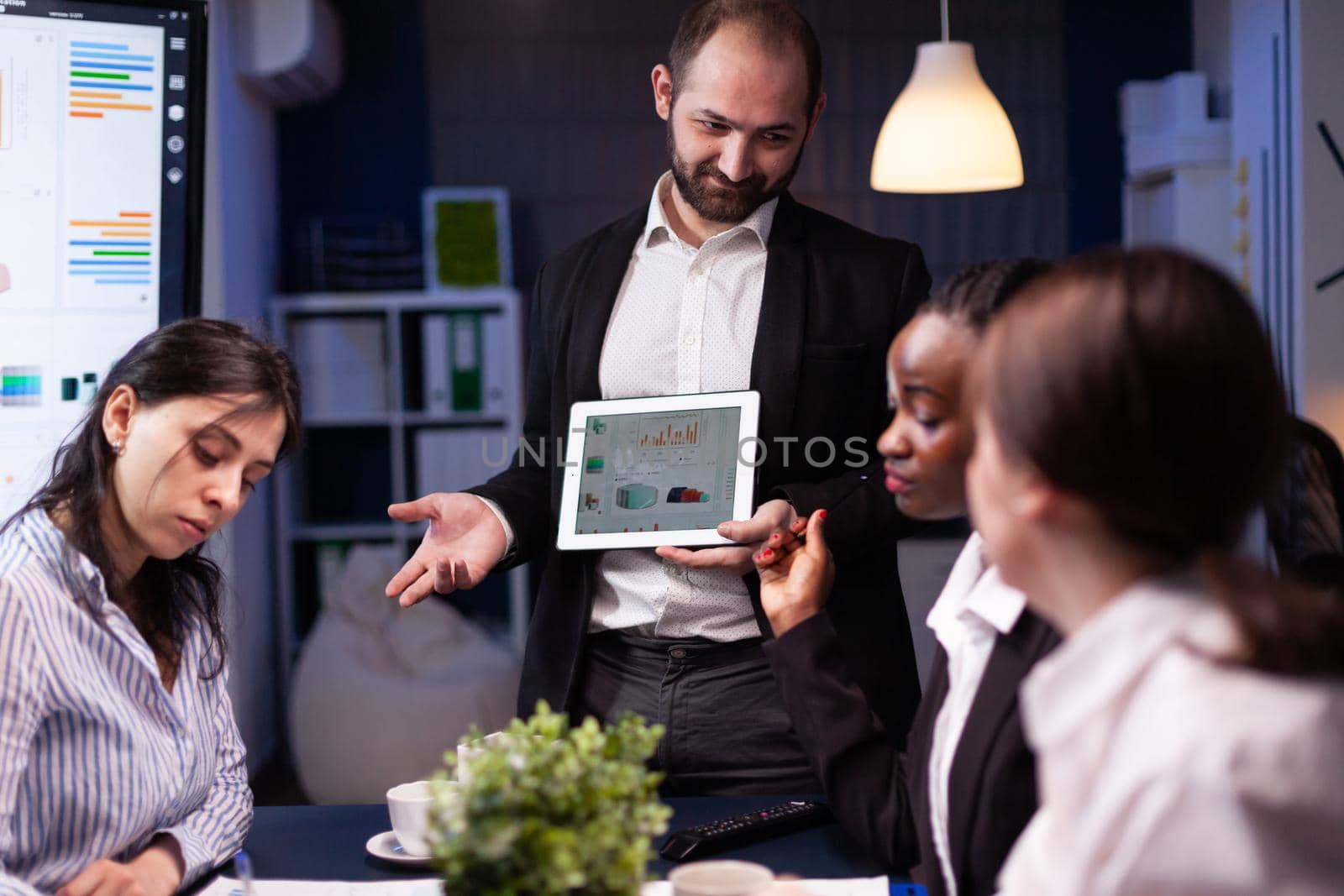 Focused workaholic entrepreneur man working overtime presenting company statistics using tablet. Diverse multi-ethnic businesspeople overworking in office meeting room late at night.