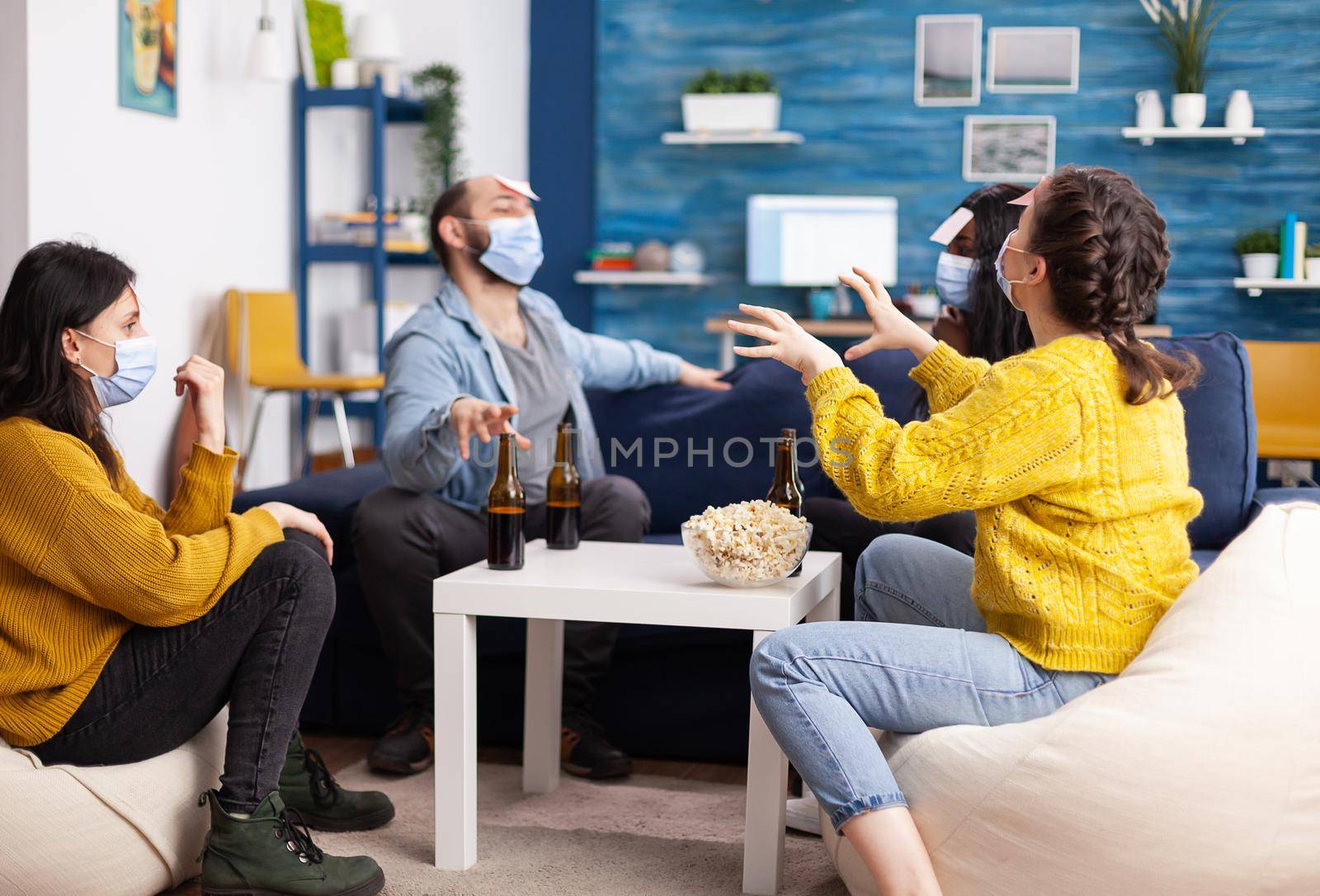 Happy cheerful multi ethnic friends playing games with sticky notes on forheads drinking beer, eating popcorn guesing keeping social distancing due to social pandemic in home living room.