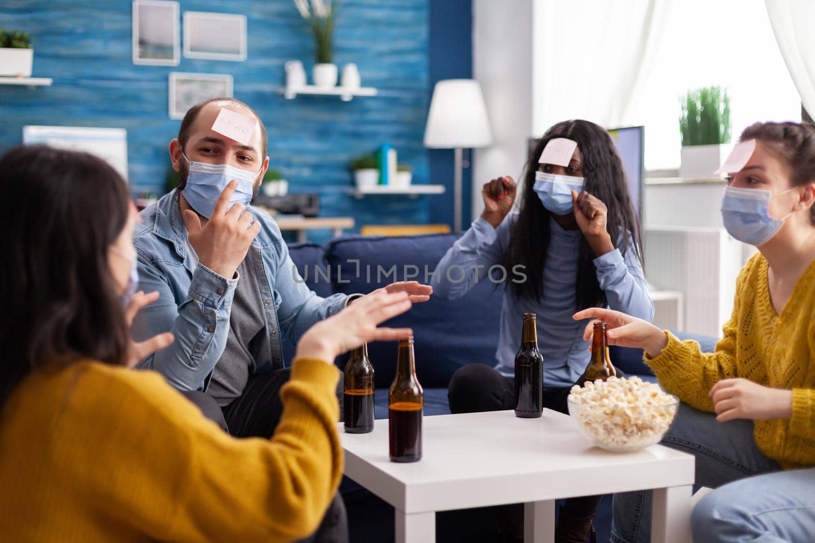 Cheerful friends with sticky notes on foreheads playing name game with beer popcron keeping social distacing with face mask to prevent spread of covid19 in home living room. Conceputal image.