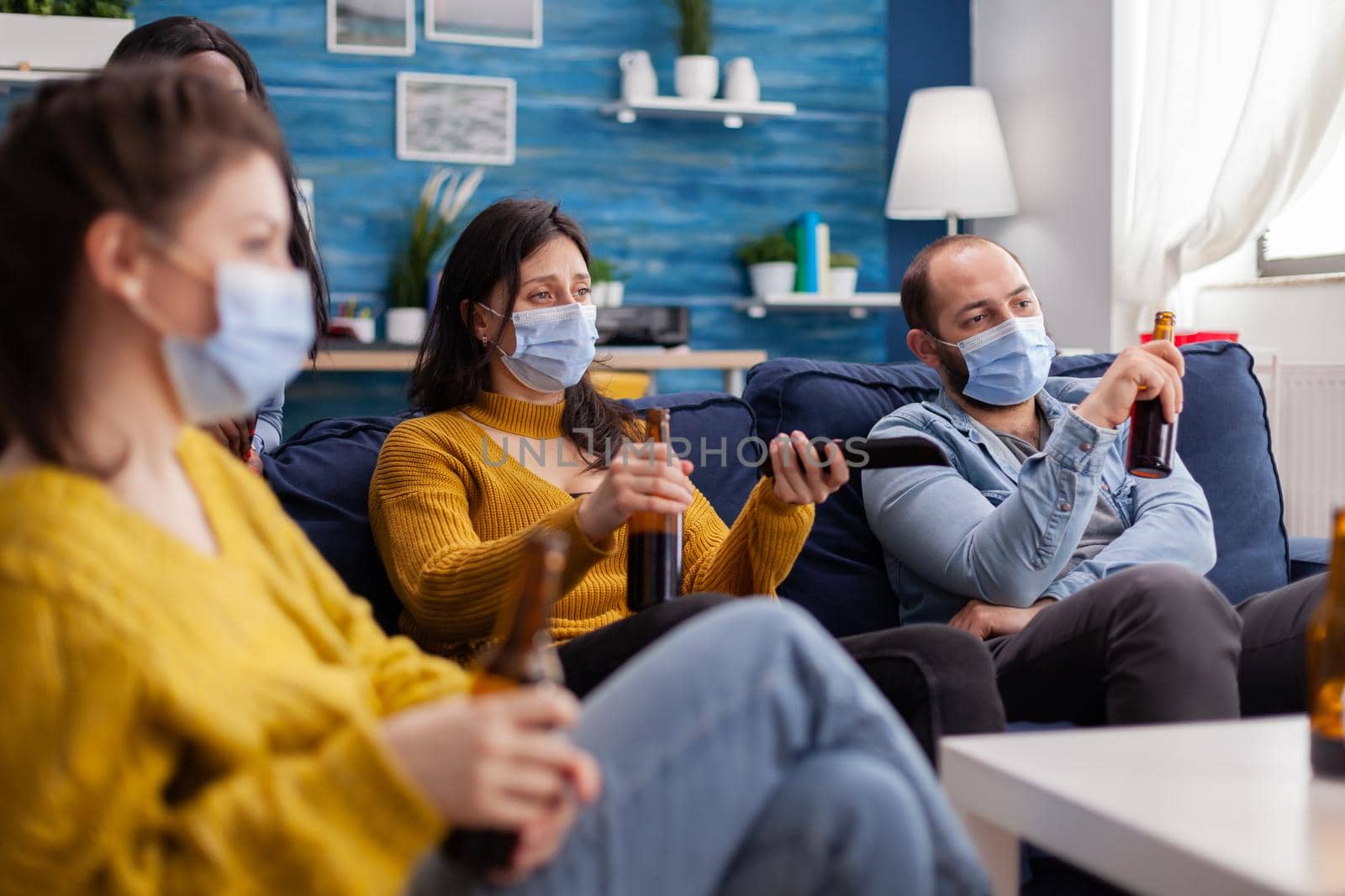 Multiethnic friends relaxing together watching tv using remote control keeping social distancing wearing face mask during coronavirus outbreak to prevent illness and sickness.
