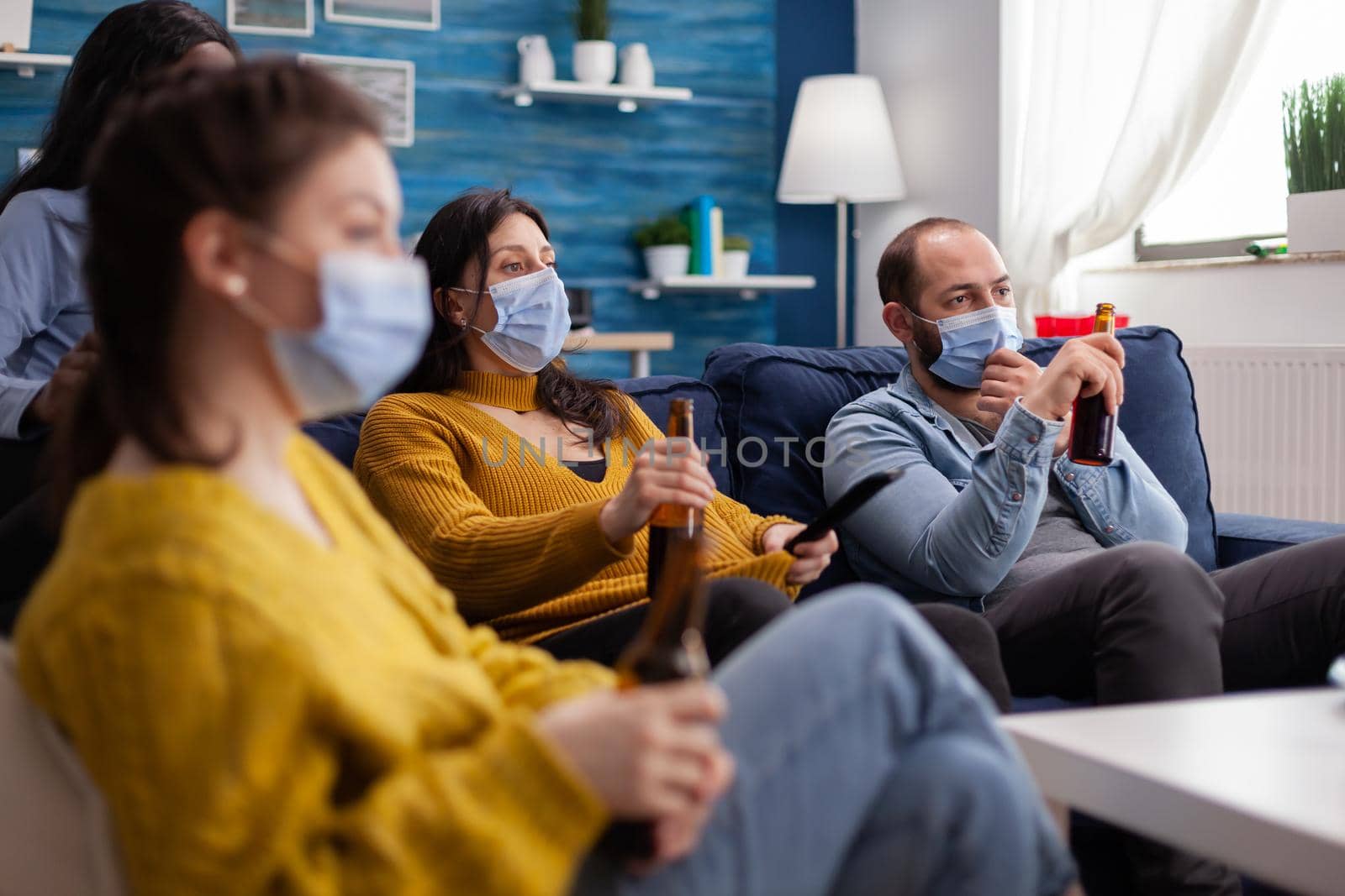 Mixed raced friends watching a film laying on couch wearing face mask to prevent infection with covid19 during social pandemic drinking beer holding remote control.