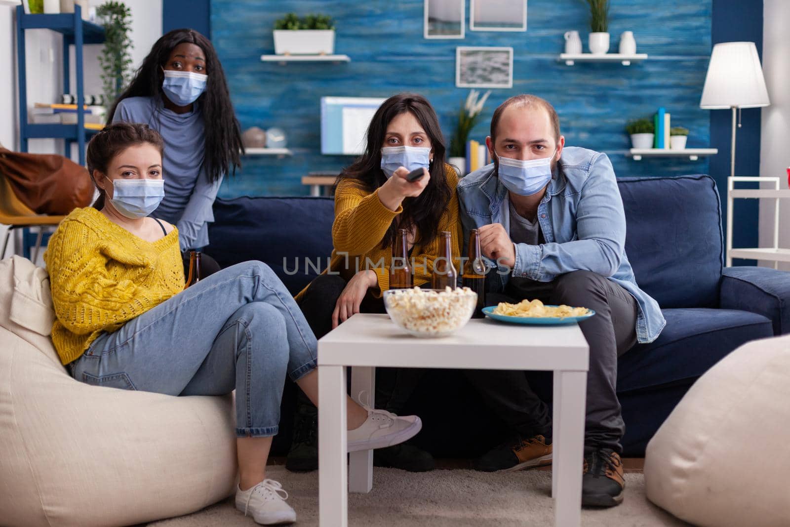 Pov of multiethnic friends using tv remote control to change channel sitting on sofa wearing face mask during covid pandemic outbreak as prevention against spread having fun.