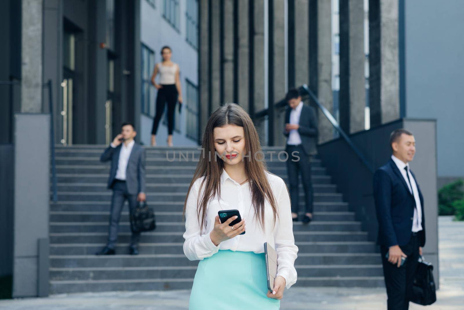 Caucasian Confident Young Business Woman is Using Smartphone App Outside near Modern Office Building by uflypro