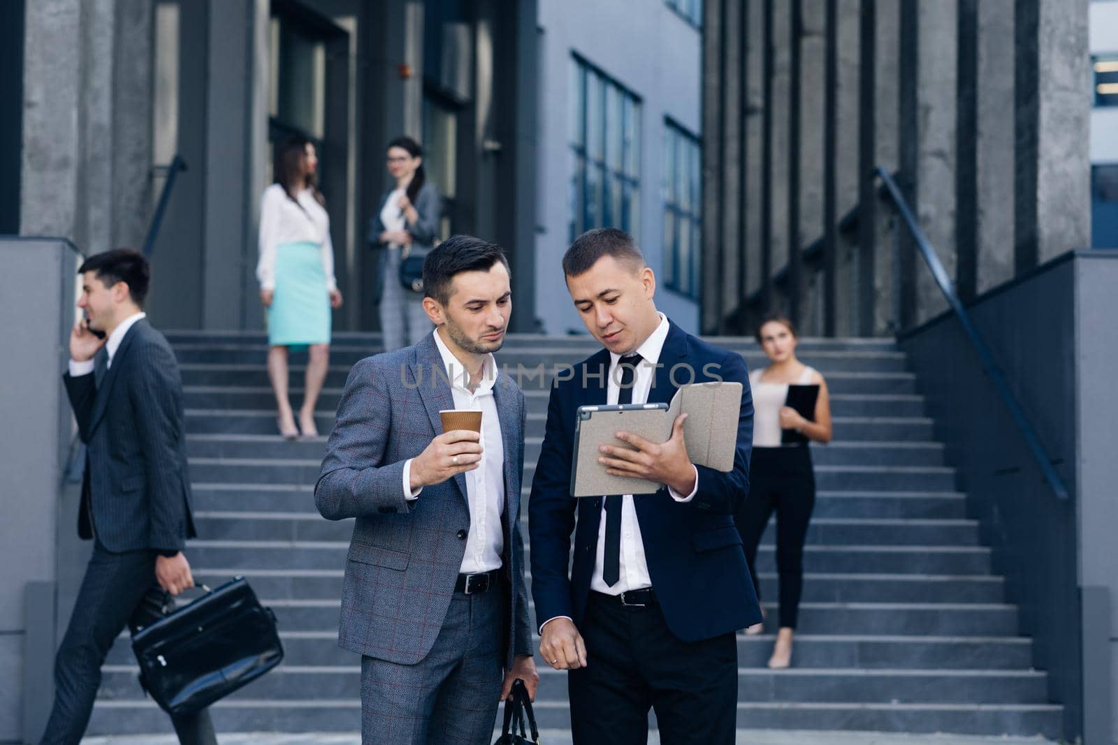 Two Caucasian handsome businessmen in suits and ties holding coffee in hands and walking from office building after work day. Stylish men talking and discussing business.