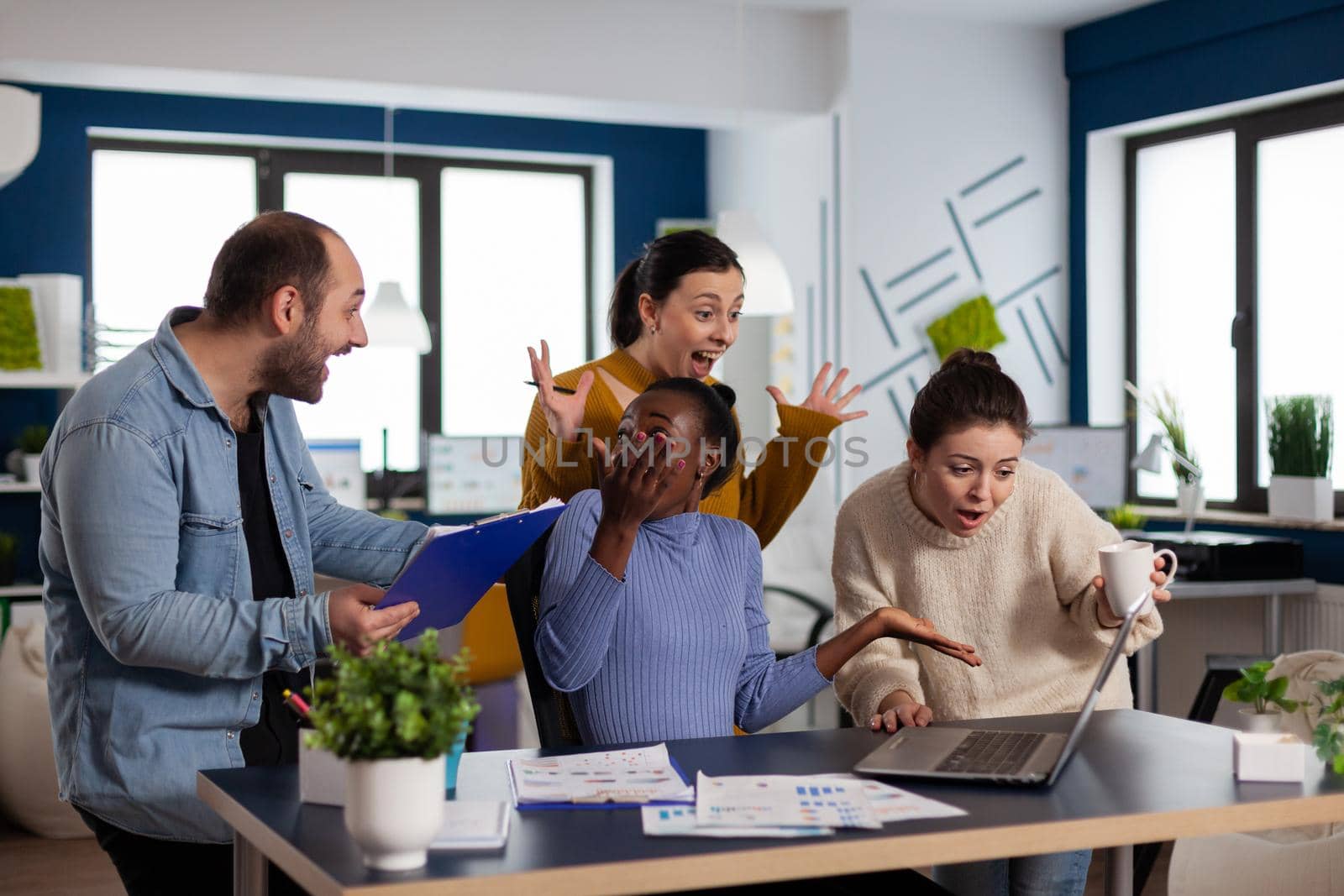 Diverse business people screaming after corporate achievement in startup office. Multiethnic coworkers business team with laptop and papers excited about project.